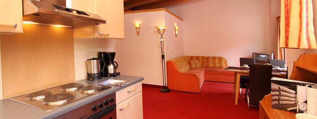 A modern kitchen with wooden shelves and an open living space. The room is cozy with a red carpet and a comfortable couch. - Haus Gondelblick - Sölden