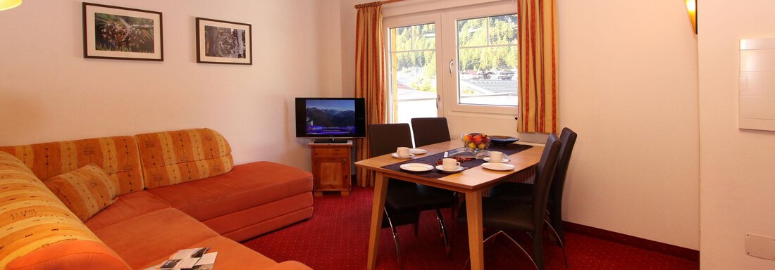 A cozy living room with an orange couch and a table. The room has a large window with a view and is nicely decorated. - Haus Gondelblick - Sölden