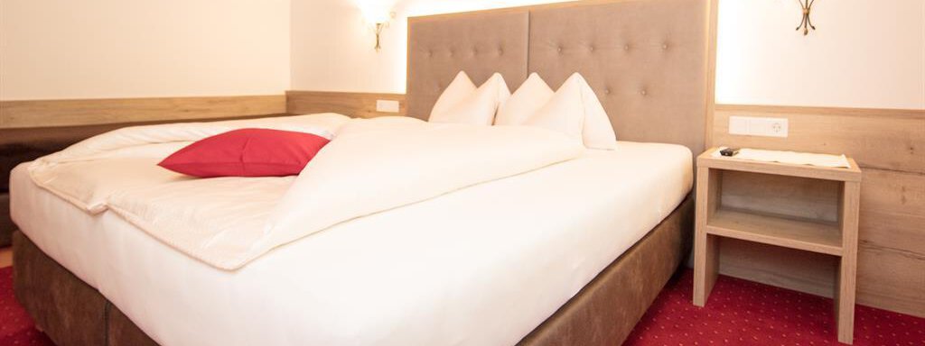 A cozy hotel room with a large double bed and red accents. A picture of a mountain landscape hangs on the wall, and the floor is carpeted with red. - Haus Gondelblick - Sölden