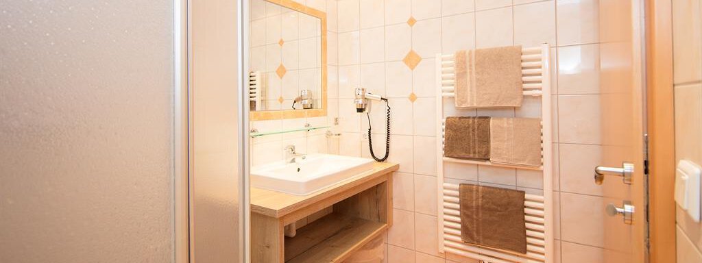 A modern bathroom with a sink and a large mirror. Towels hang on a towel warmer and the floor is tiled with beige tiles. - Haus Gondelblick - Sölden