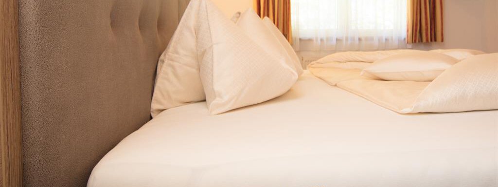 A cozy bedroom with a comfortable bed and pillows. The room has large windows with curtains and a wooden little table. - Haus Gondelblick - Sölden