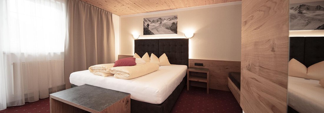 A cozy hotel room with a large bed and wooden furniture. The room is bright and well-lit, with large windows and simple, modern decor. - Haus Gondelblick - Sölden