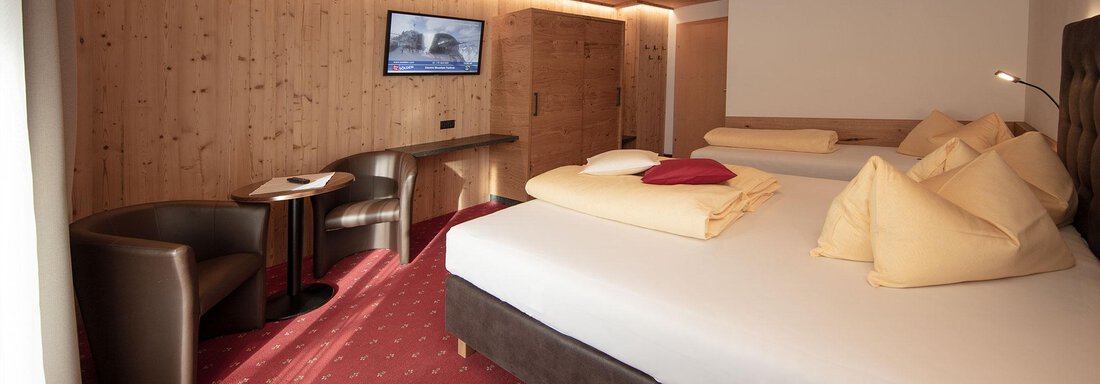 A modern hotel room with wooden walls and a red carpet. There are two beds, a television, and a small table with chairs. - Haus Gondelblick - Sölden
