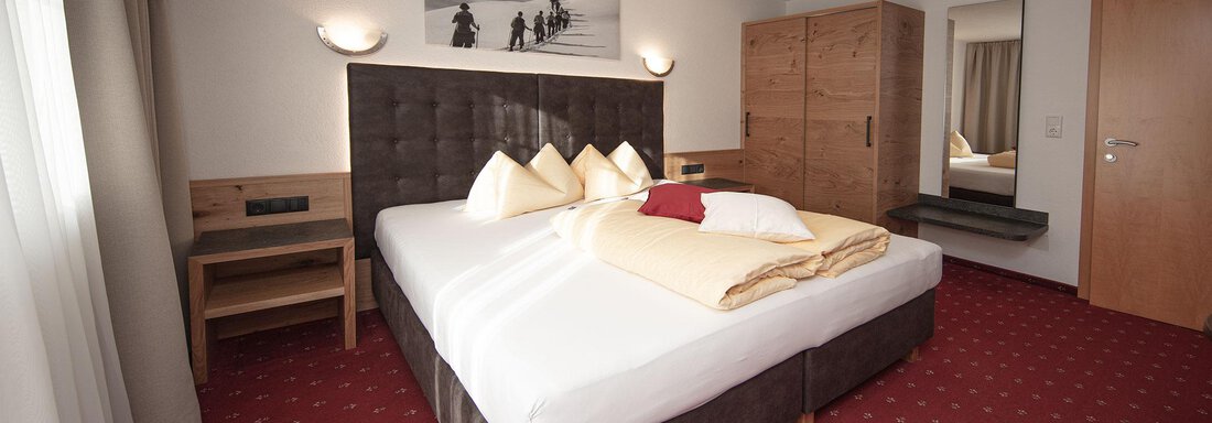 A modern hotel room with a large double bed and beige bedding. The walls are light, and the furniture is made of wood with a cozy carpet. - Haus Gondelblick - Sölden
