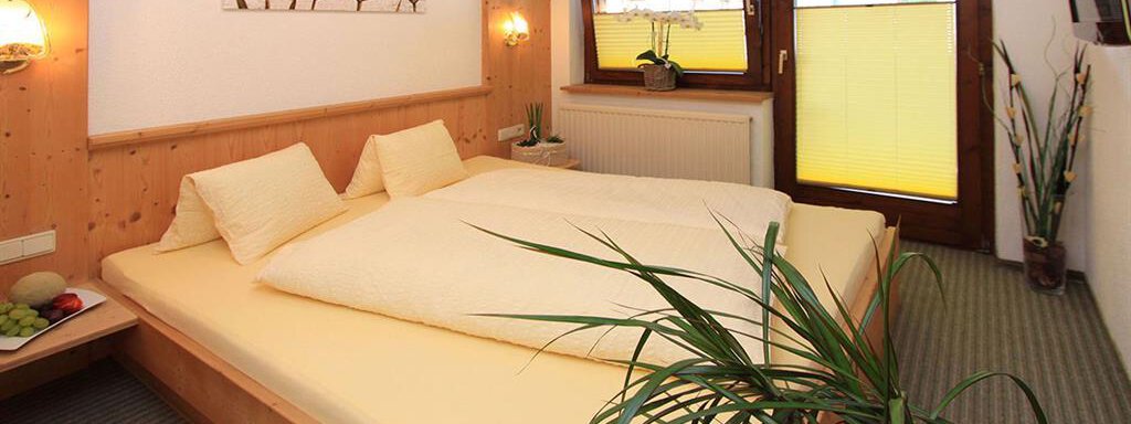 A cozy bedroom with a large bed and wooden details. The windows are covered with yellow blinds, and some plants create a pleasant atmosphere. - Haus Ingrid - Sölden