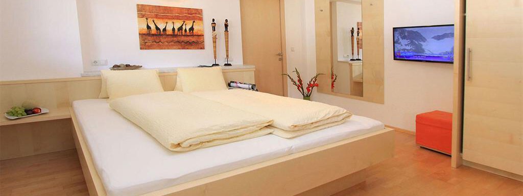 A modern bedroom with a large bed and light wood decor. Decorations hang on the wall, and a television is visible. - Haus Ingrid - Sölden