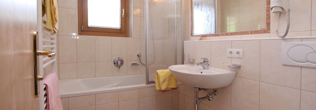 A bright bathroom with a bathtub, sink, and shower. The walls are tiled and there is a window for natural light. - Haus Montjola - Sölden