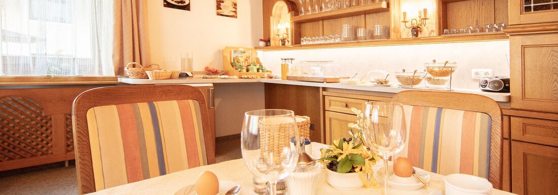 An inviting breakfast table set with dishes and a fresh bouquet of flowers. In the background, a bright kitchen with wooden shelves and dishware is visible. - Haus Montjola - Sölden