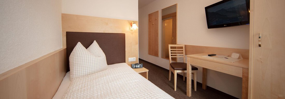 A simple single room with a cozy bed and a wooden table. A television hangs on the wall, and the room features a cohesive, modern design. - Haus Montjola - Sölden