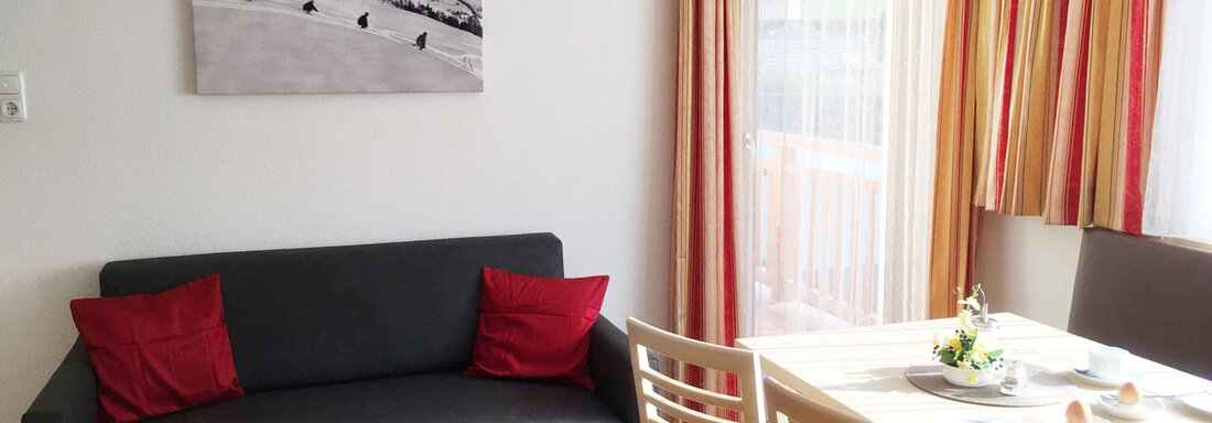 A cozy living area with a gray sofa and red cushions. On the table, there are dishes and a small floral decoration. - Haus Montjola - Sölden