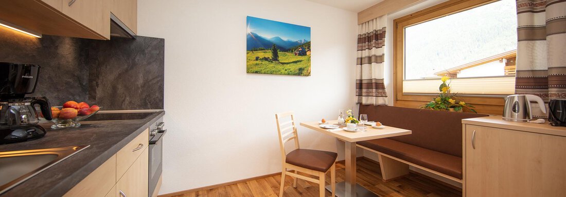 A modern kitchen with hardwood flooring and light cabinets. There is a dining area with a table and a bench, as well as a window overlooking the outdoors. - Haus Montjola - Sölden