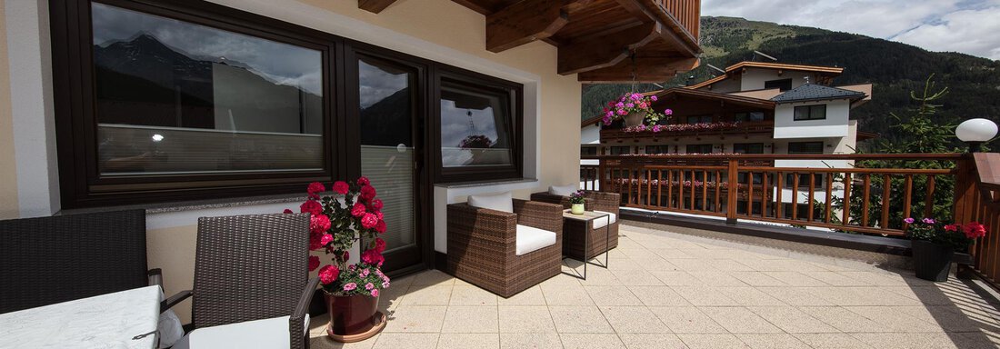A cozy terrace with seating and blooming plants. In the background, mountains and more houses are visible. - Haus Montjola - Sölden