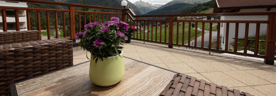 A balcony with a view of the mountains and a green landscape. On the table is a blooming plant in a light pot. - Haus Montjola - Sölden