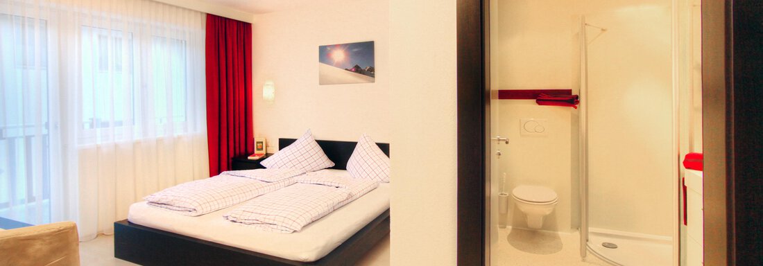 A modern room with a double bed, a chair, and large windows. In the background, there is a private bathroom with a shower. - Haus PIA by A CASA - Sölden