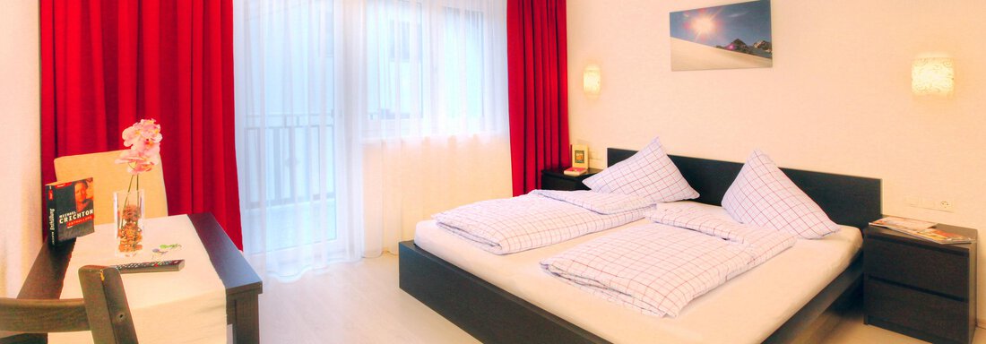 A modern bedroom with a large bed, red curtains, and a small table. The room feels bright and friendly. - Haus PIA by A CASA - Sölden