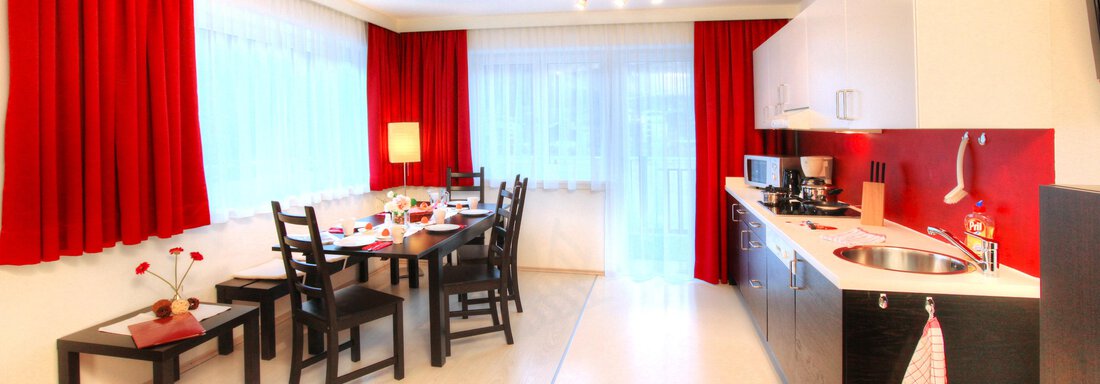 A modern kitchen with a dining table and chairs. Red curtains and a well-equipped kitchenette create an inviting atmosphere. - Haus PIA by A CASA - Sölden