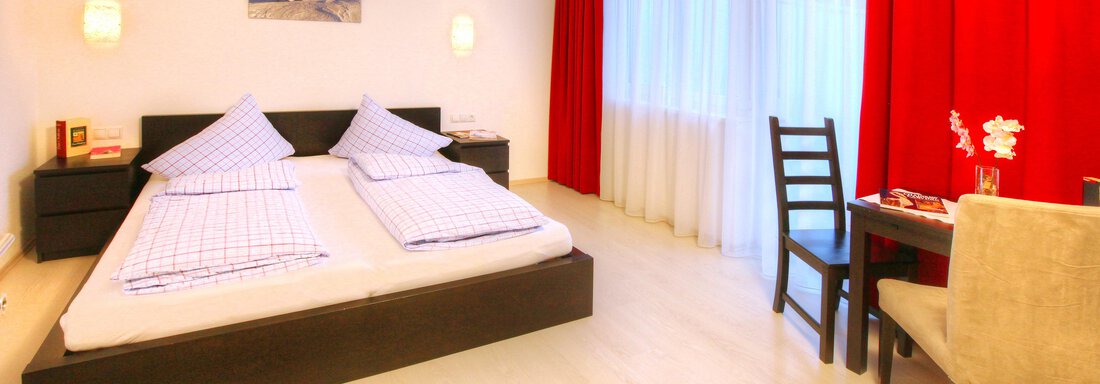 A modern bedroom with a double bed and white bedspreads. On one wall there are red curtains and a small table with chairs. - Haus PIA by A CASA - Sölden