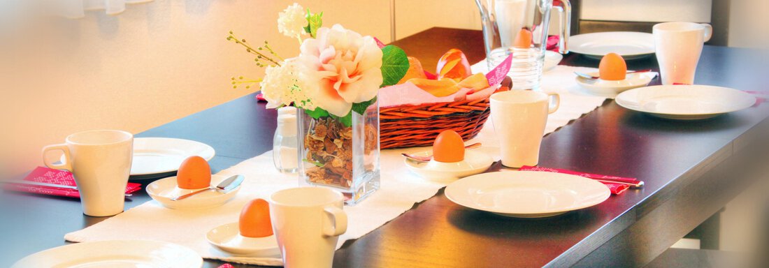 A beautifully set table with white plates, cups, and orange eggs. In the center is a flower vase and a basket. - Haus PIA by A CASA - Sölden
