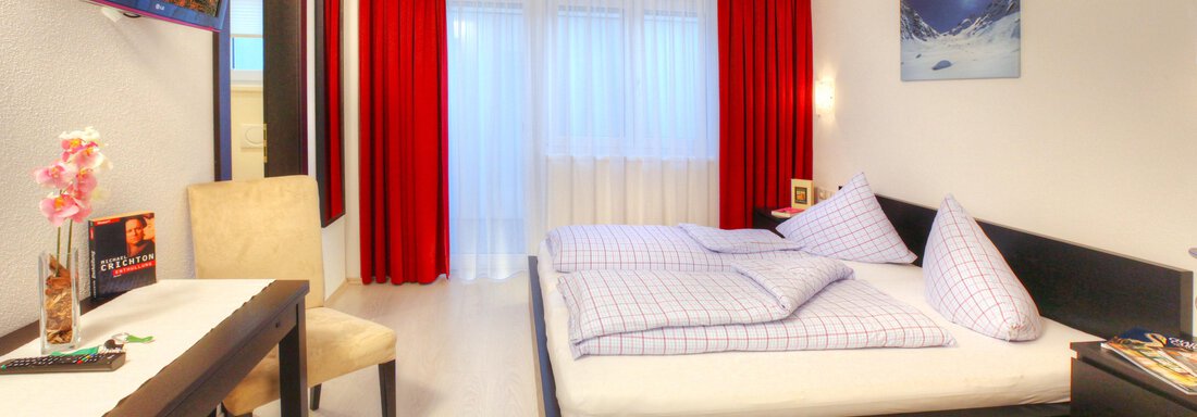 A modern hotel room with a double bed and a desk. Bright walls and red curtains create an inviting atmosphere. - Haus PIA by A CASA - Sölden