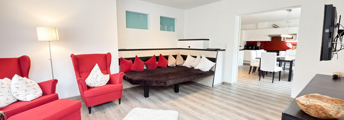 A modern living room with two red chairs and a cozy, wavy couch. The room features a light wooden floor and an open connection to the kitchen. - Haus PIA by A CASA - Sölden