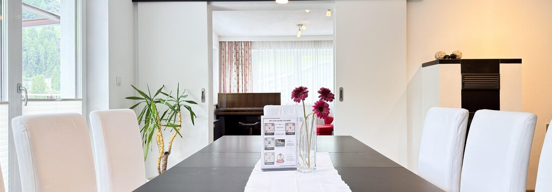 An elegant dining area with a large, dark table and white chairs. In the background, there are windows and a plant visible, bringing brightness. - Haus PIA by A CASA - Sölden