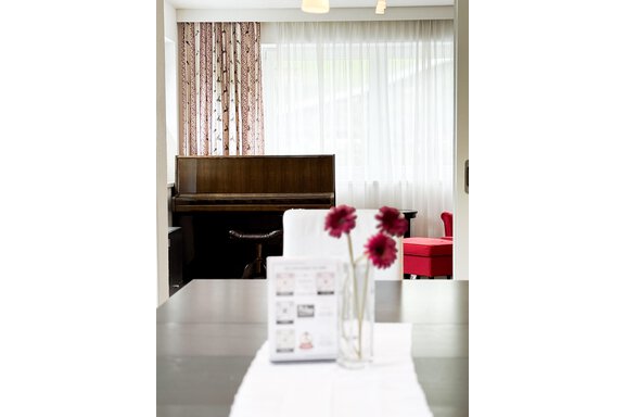 A cozy room with a piano in the background and a table with a vase full of flowers in the foreground. Bright curtains let plenty of light in. - Haus PIA by A CASA - Sölden