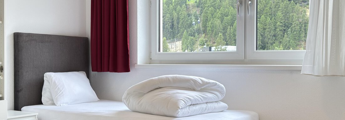 A simple bedroom with a single bed and white bed linen. Through the window, a green mountain landscape is visible. - Haus PIA by A CASA - Sölden