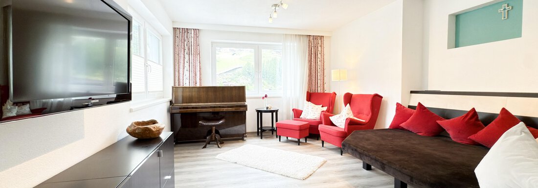 A modern living room with a large TV and a cozy sofa. Red accent pillows and a wooden chair give the room an inviting atmosphere. - Haus PIA by A CASA - Sölden