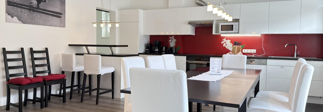 A modern dining room with a large table and white chairs. The walls are light, and the kitchen unit has red accents. - Haus PIA by A CASA - Sölden