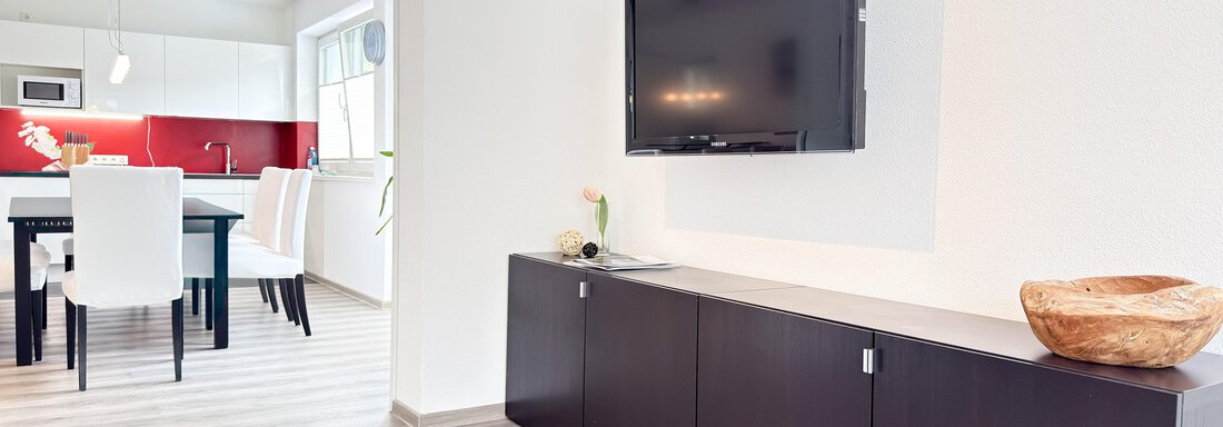 A modern living room with a flat-screen TV on the wall. In the background, a bright kitchen with a dining table and chairs can be seen. - Haus PIA by A CASA - Sölden