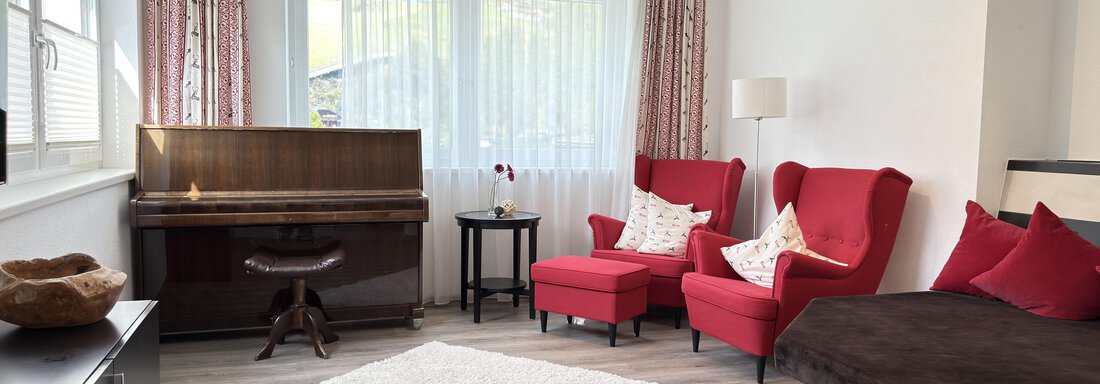 A cozy living room with two red armchairs and a piano. The room has large windows that let in plenty of light, and a bright carpet on the floor. - Haus PIA by A CASA - Sölden