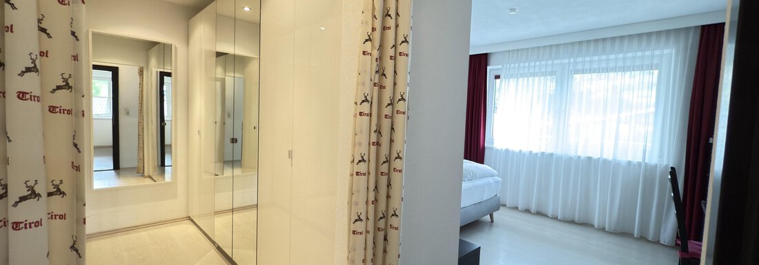 A bright, modern room with a large mirror and a bedroom in the background. The windows are covered with white curtains, allowing plenty of daylight in. - Haus PIA by A CASA - Sölden