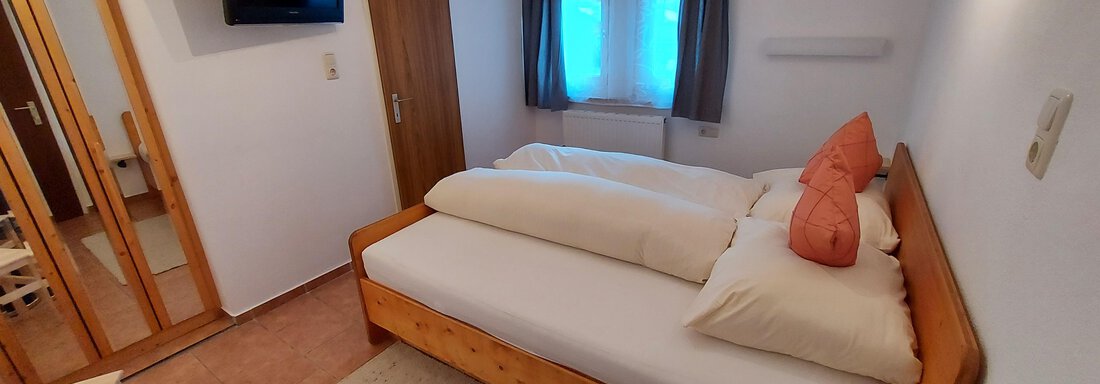 A small, cozy bedroom with two beds and orange pillows. There is a television, a desk, and a large mirror. - Haus Pitzwald - Sölden