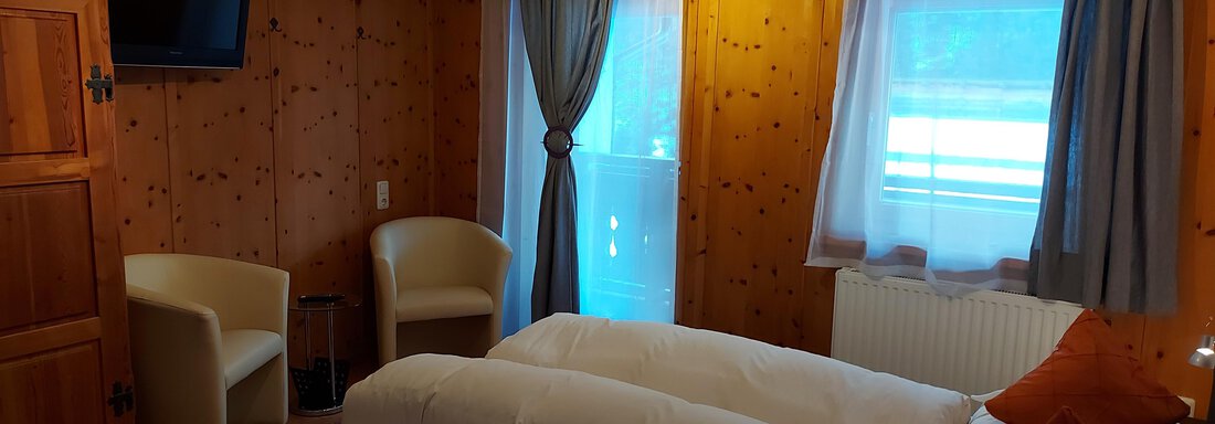 A cozy hotel room with two beds, a television, and a small seating area. The walls have a wood look and the windows let in plenty of natural light. - Haus Pitzwald - Sölden