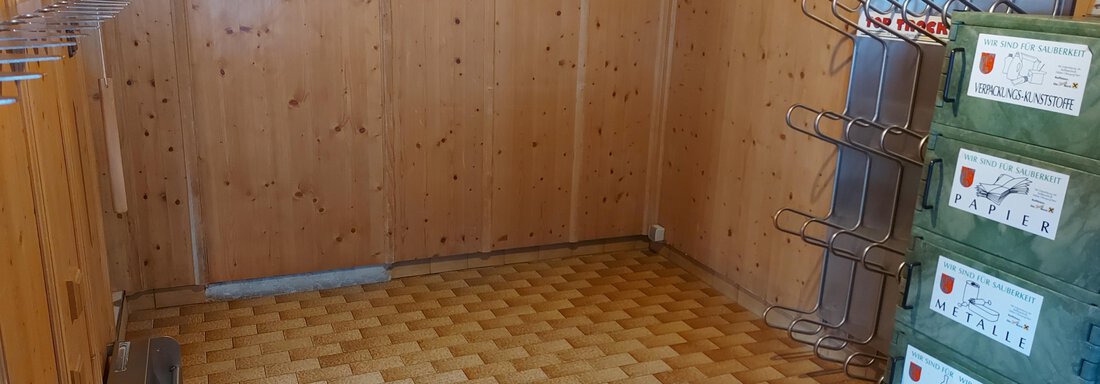 An empty room with wooden paneling and a window. Clothes hooks hang on the walls, and there are green boxes in one corner. - Haus Pitzwald - Sölden