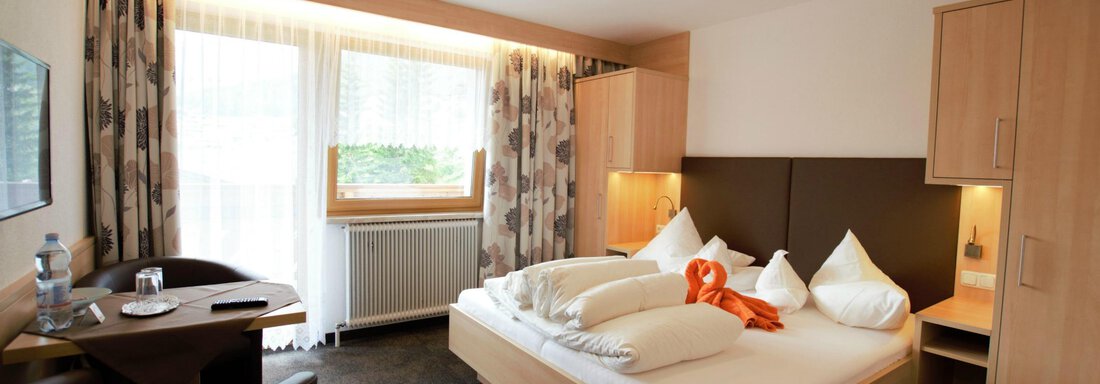 Double room south | © Schneider Eva A cozy room with a double bed, two pillows, and orange decor. There is a small table, chairs, and large windows that let in plenty of light. - Haus Rehwinkl - Sölden