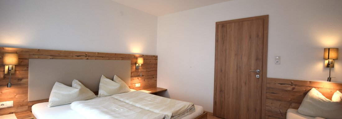 A modern bedroom with a large bed and wooden furniture. The walls are light, and there is warm light from the wall lamps. - Haus Romana - Sölden