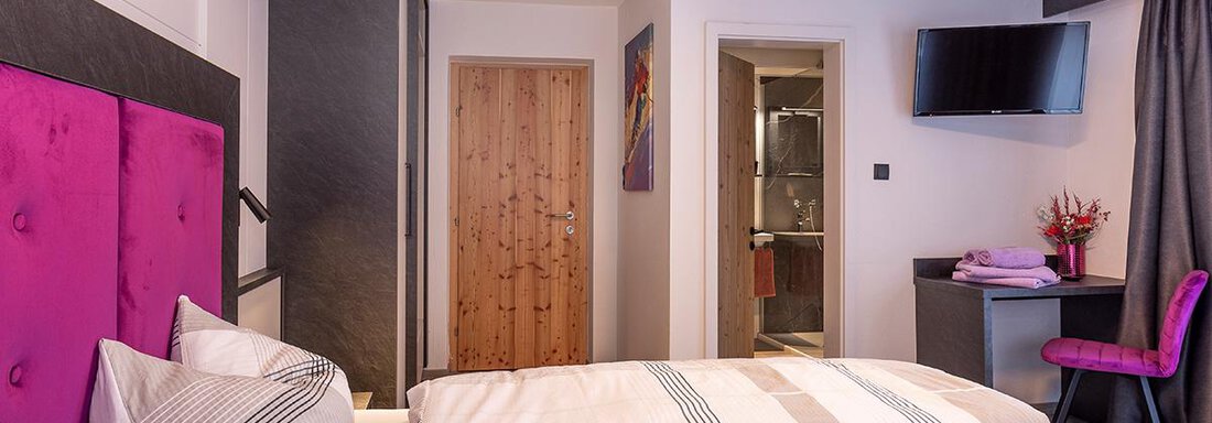 A modern bedroom with two beds and a purple headboard wall. In the background, a television and a bathroom can be seen. - Haus Sieglinde Color - Sölden