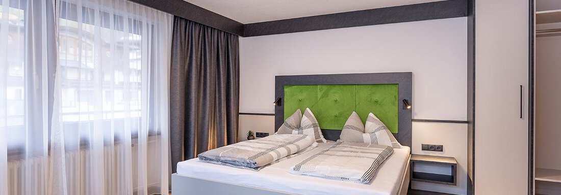 A modern bedroom with a double bed and an elegant green accent wall. Large windows provide plenty of daylight and a pleasant atmosphere. - Haus Sieglinde Color - Sölden