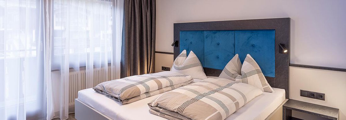 Doppelzimmer Sieglinde Color Sölden | © Sieglinde Color Sölden (CC C) A modern bedroom with a large bed and blue accents. There are curtains and a bright, inviting atmosphere. - Haus Sieglinde Color - Sölden