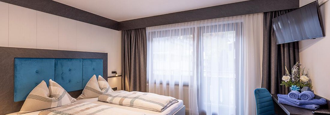 A modern bedroom with a large double bed and blue pillows. The room has large windows with curtains and a desk in a tranquil atmosphere. - Haus Sieglinde Color - Sölden