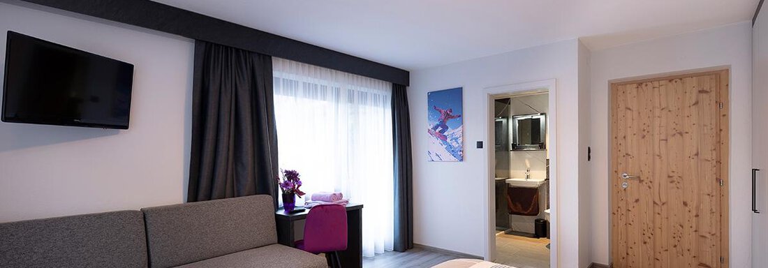 A modern hotel room with two beds and a cozy sitting area. Daylight comes through the window and the bathroom is visible in the background. - Haus Sieglinde Color - Sölden