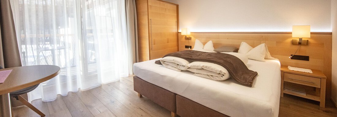 A modern hotel room with a large bed and wooden floor. The floor-to-ceiling windows provide plenty of light and a view of a balcony. - Haus Sonnwend - Sölden