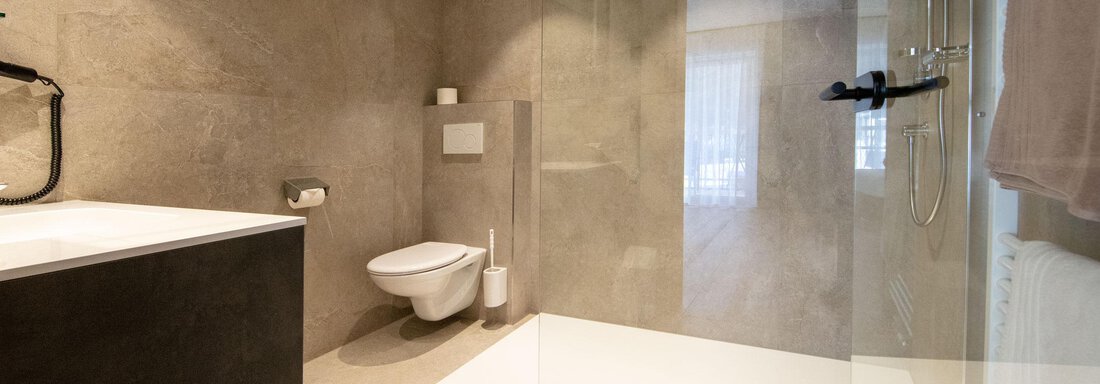 A modern bathroom with a clear shower and a freestanding bathtub. The walls are in neutral gray tones, and there is an elegant rug. - Haus Sonnwend - Sölden