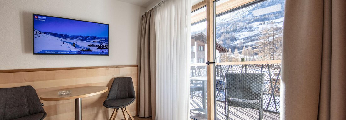 A modern room with a TV, a table, and two chairs. The windows open to a balcony with a view of the mountains. - Haus Sonnwend - Sölden