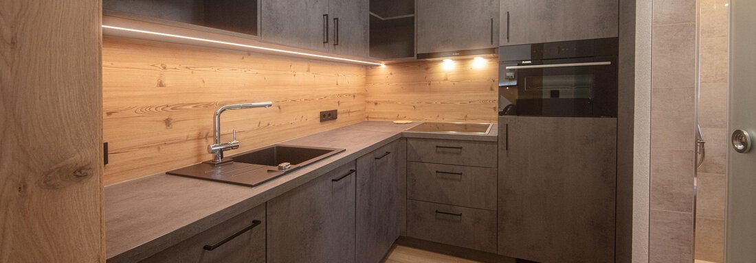 A modern kitchen with gray cabinets and wood paneling. It features a sink, a countertop, and integrated lighting. - Haus Sonnwend - Sölden