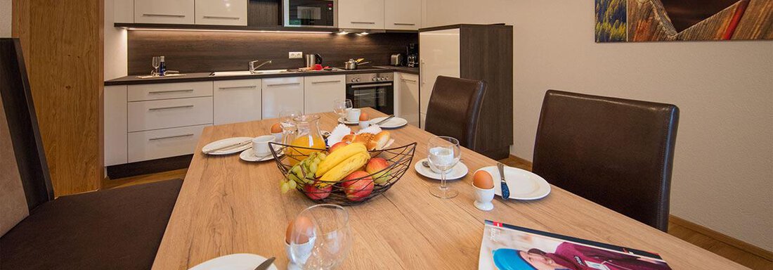 A modern kitchen with a dining area. The table is set with fruit, dishes, and a magazine. - Haus Stefanie - Sölden