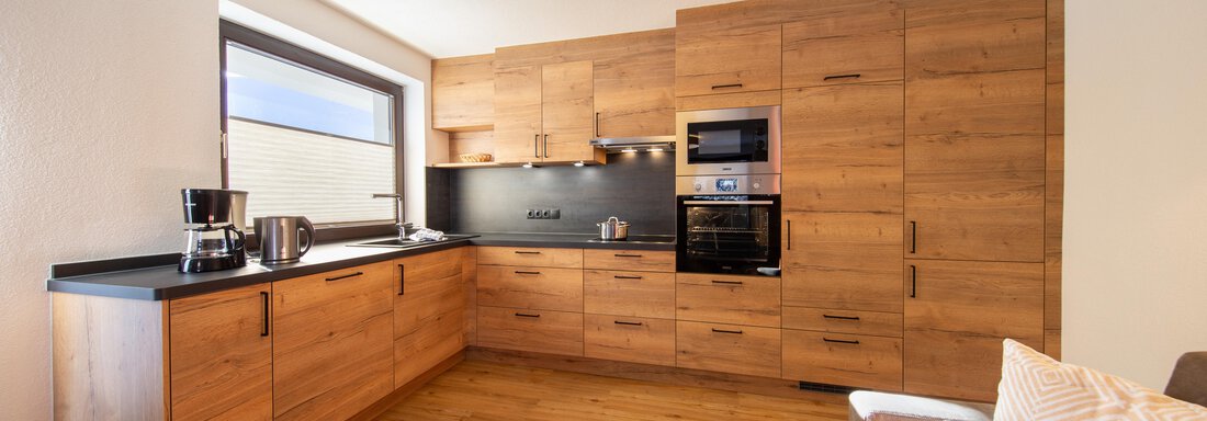 A modern kitchen with wooden furniture and a black workspace. There is a large window area and a bright ceiling light. - Haus Stefanie - Sölden