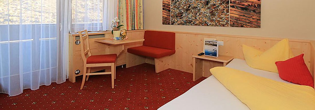 Einzelzimmer mit Balkon A cozy room with wooden furniture and a red carpet. There is a window with curtains and a table with a chair. - Haus Stefanie - Sölden