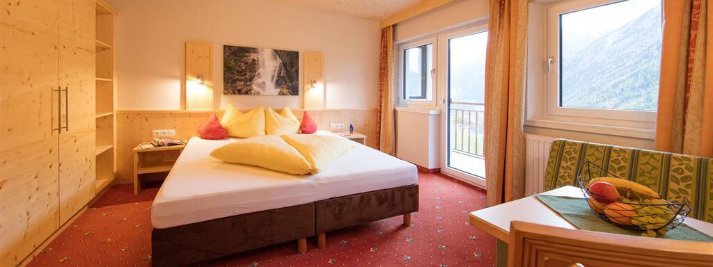A cozy bedroom with a large bed and colorful pillows. There is a table with fruit and windows with a view of the mountains. - Haus Stefanie - Sölden